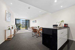 A modern kitchen with a black countertop, a dining area with a round table, and a view of a building through large windows. - Manhattan CBD Stay, Resort-Style Prime Location (City)