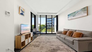Een moderne woonkamer met een bank, televisie, houten kast en een groot raam met uitzicht op gebouwen en groen.