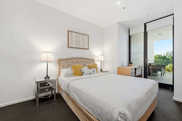 Ein modernes Hotelzimmer mit einem großen Bett, zwei Nachttischen, einem kleinen Schreibtisch, einem Sessel und einem Balkon mit Glasdoor.