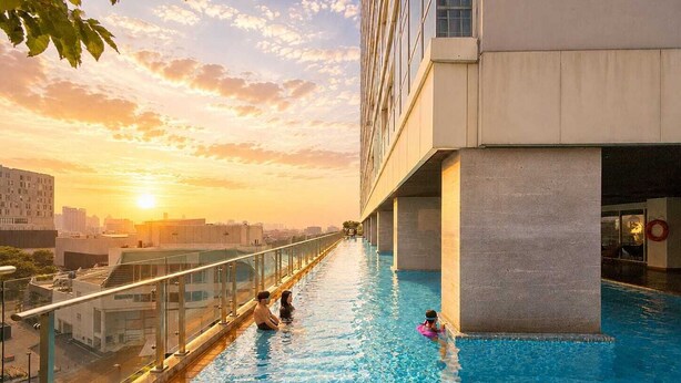 Piscina no terraço com guarda-corpos de vidro, vista desobstruída da cidade e um pôr do sol ao fundo.