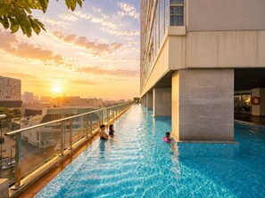 Une piscine sur le toit, entourée de rampes de verre, offrant une vue dégagée sur le paysage urbain, avec un coucher de soleil en toile de fond.