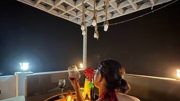 A woman seated at a table with a bottle of wine, a glass, and a candle. The setting is indoors with a ceiling structure and string lights.