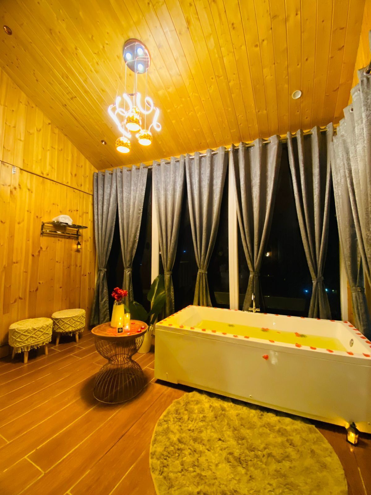 Una habitación con techo de madera, una bañera grande con pétalos de rosa, un tapete redondo y una pequeña mesa con un jarrón de flores.