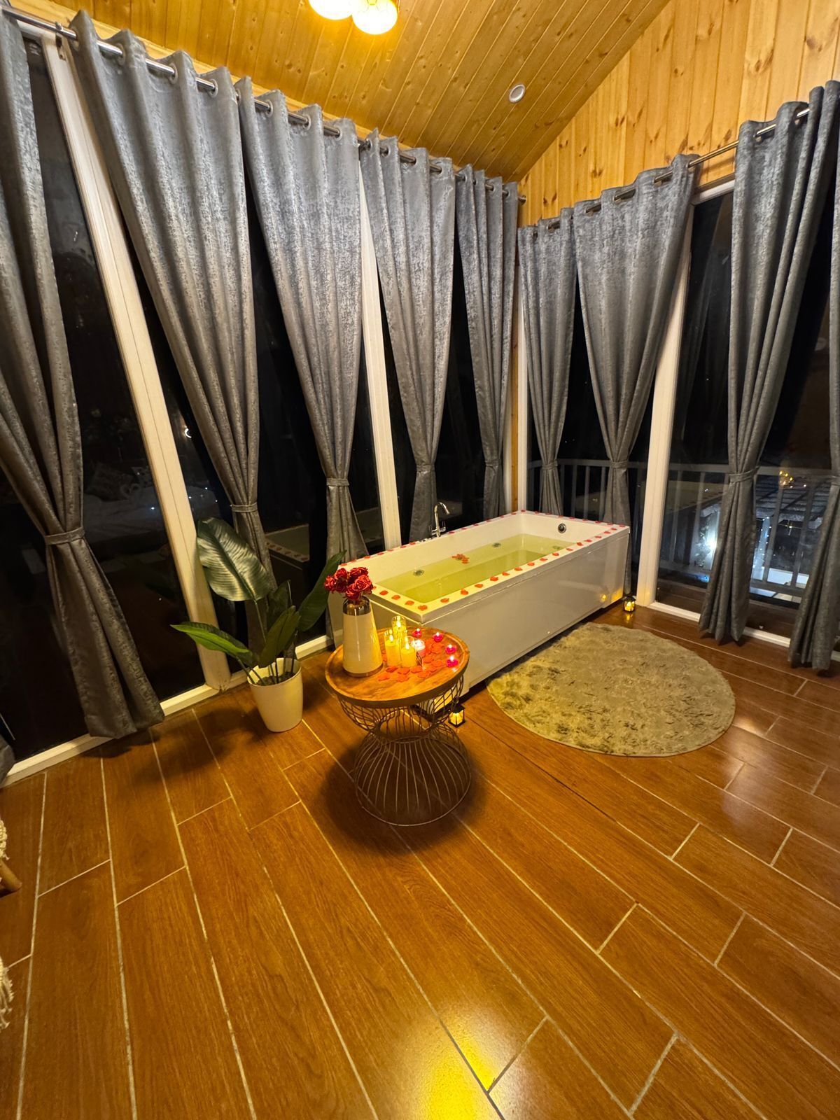 A room with a bathtub, a potted plant, a small table with candles, and a round rug.
