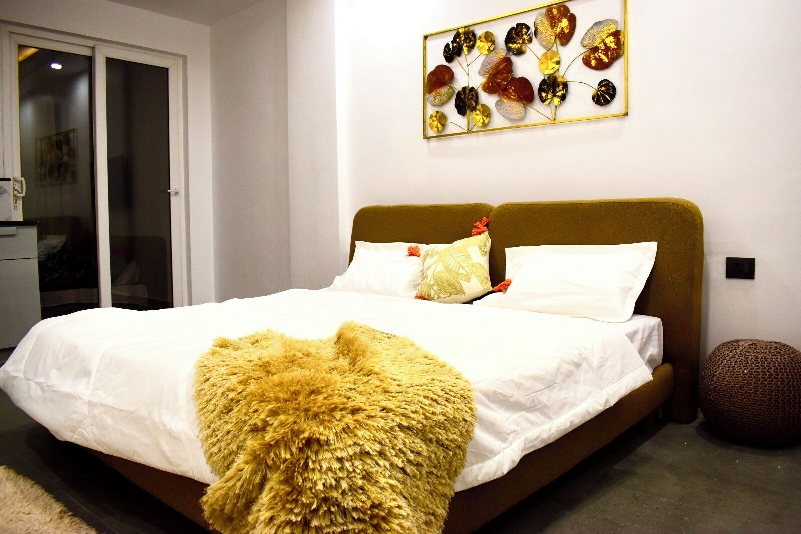 Een slaapkamer met een bed, kussens, een zachte plaid, een kunstwerk aan de muur en een klein rond poef.
