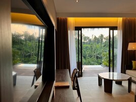 Habitación de hotel moderna con un ventanal grande que ofrece vistas a exuberante vegetación. Hay una mesa de madera, un sofá y una silla en la habitación.