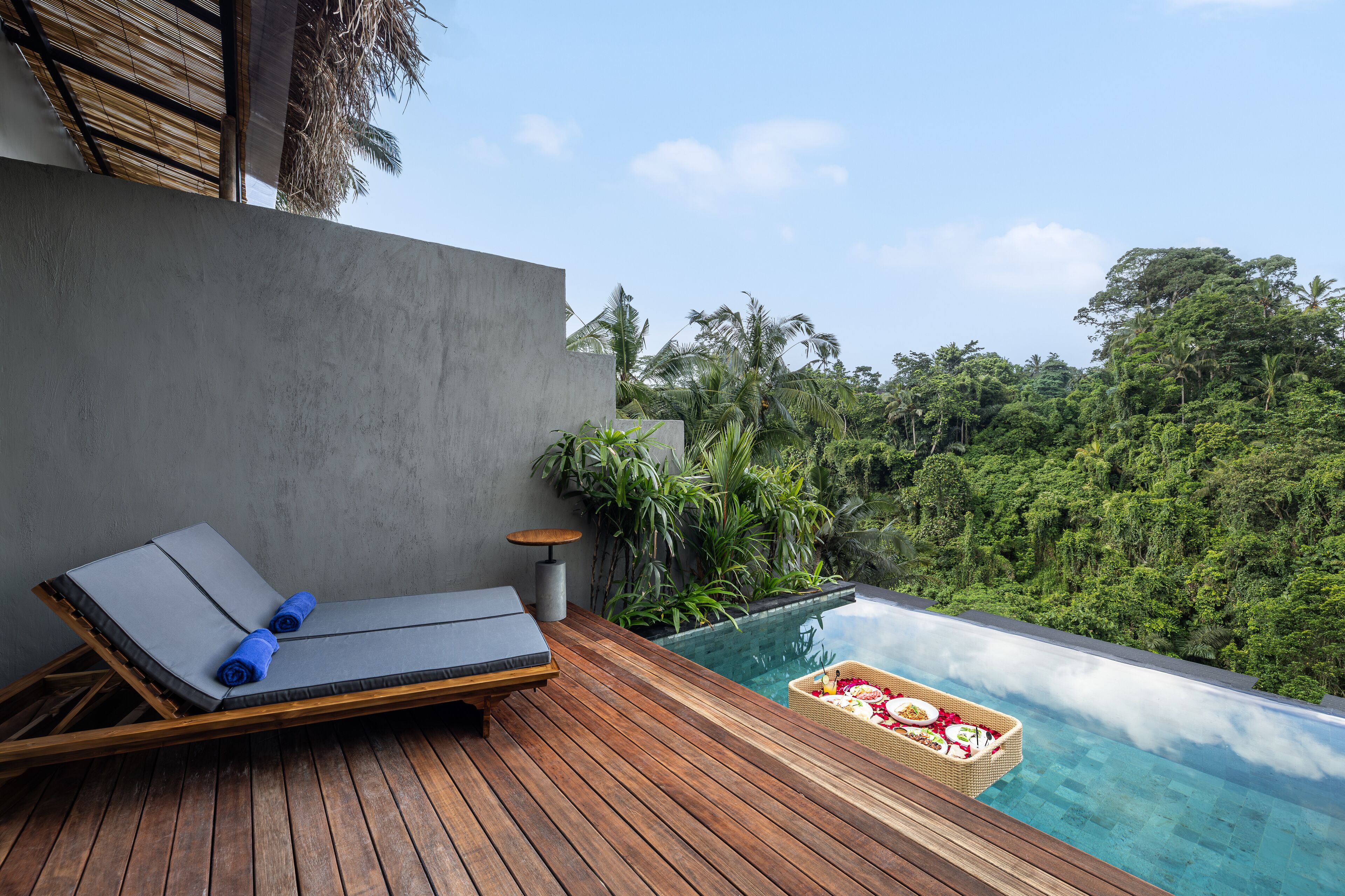 A wooden deck with a lounge chair and a small table, overlooking a clear pool with a tropical view.