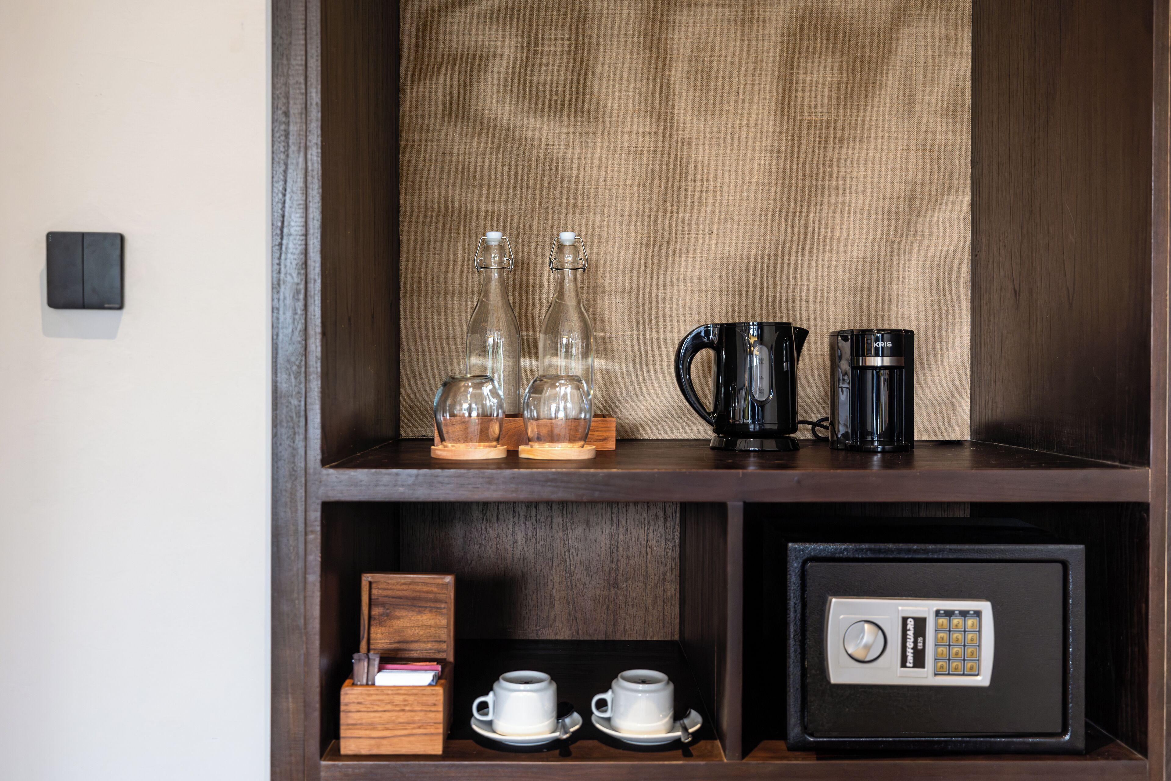 A wooden shelf with glass bottles, a black coffee maker, a safe, and two white cups.