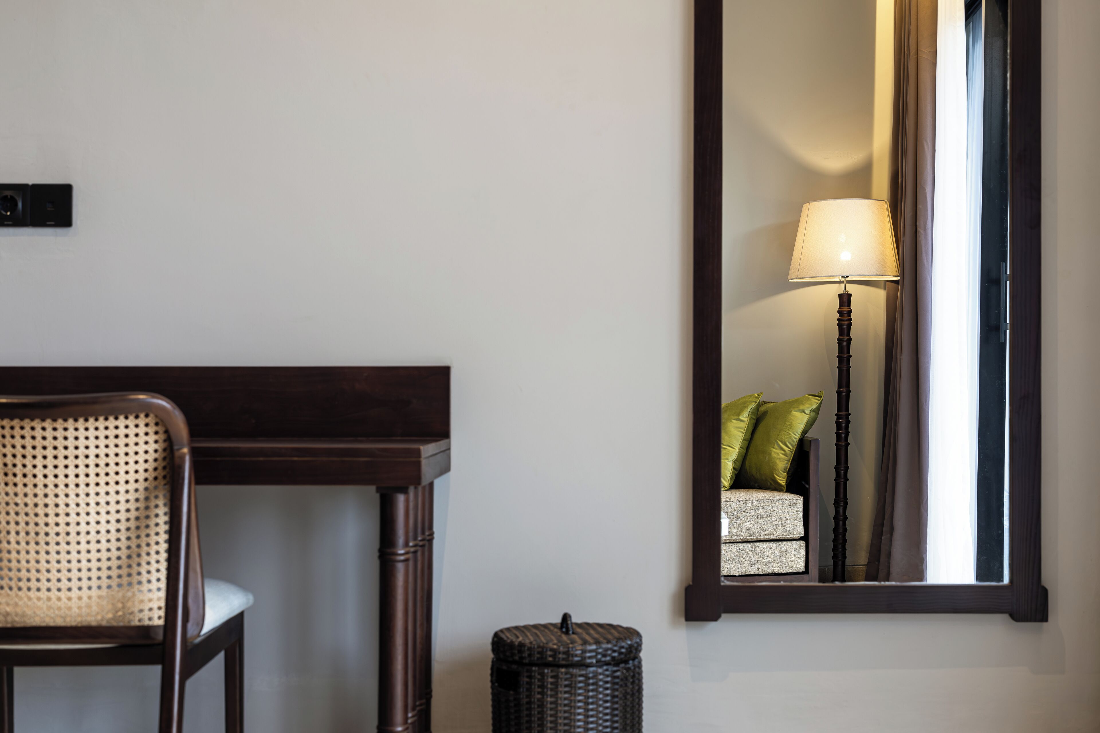 A room with a wooden chair, a small table, a lamp, a mirror, and a wicker basket.