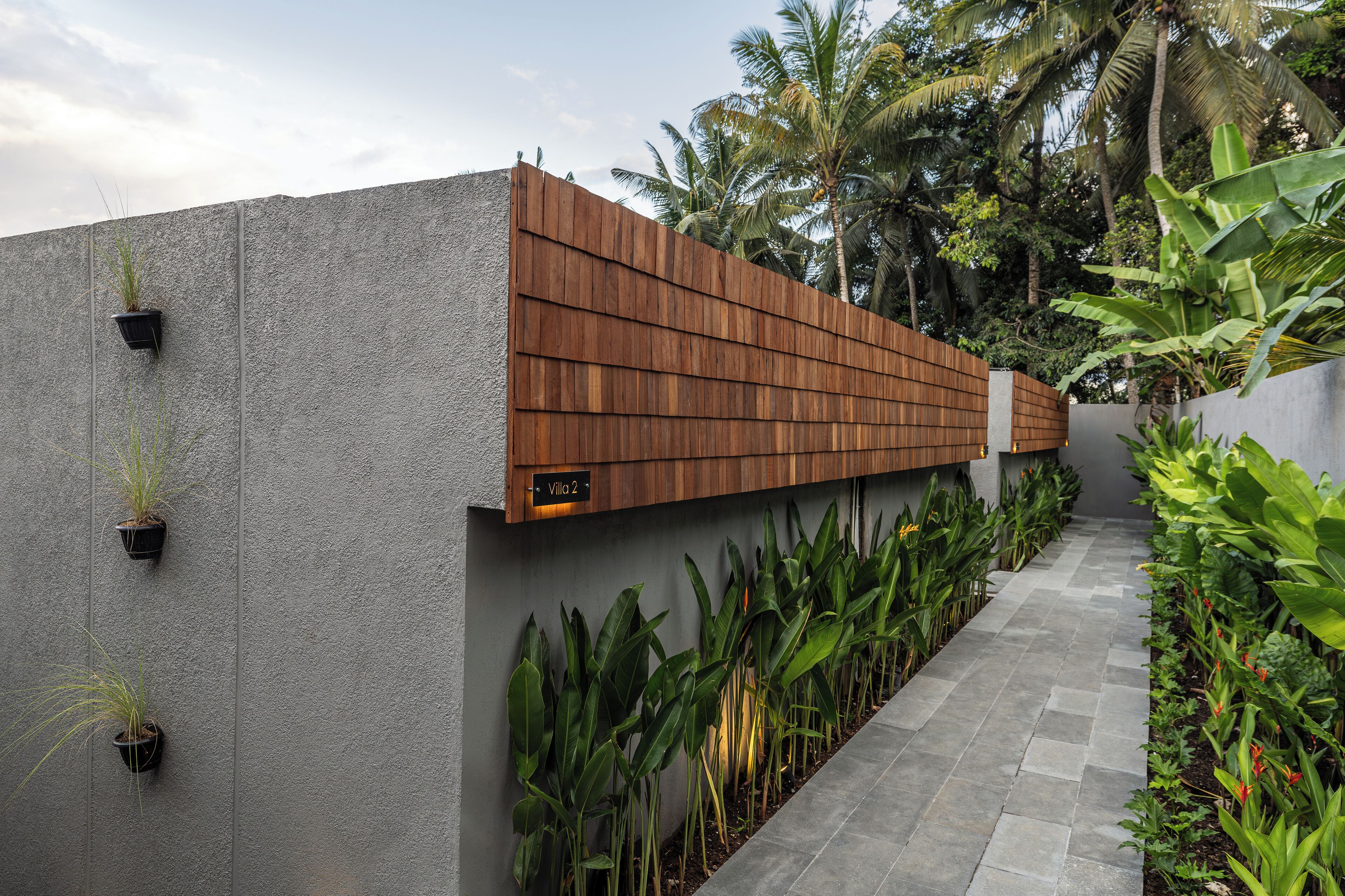 A modern outdoor pathway with a wooden fence, potted plants, and a sign indicating 'Villa 2'.