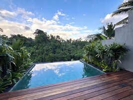 Una terraza de madera que conduce a una piscina infinita rectangular con vistas a un bosque frondoso y verde.