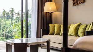 A modern hotel room with a sofa, a table, and a lamp. There is a view of greenery outside the window. - The Clifton Villas Ubud (Ubud)