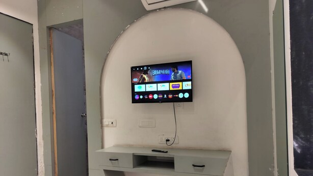 A room with a TV mounted on a curved wall, a desk with drawers, and a bed with a red blanket. - ShrI Balaji Dham (Vrindavan)