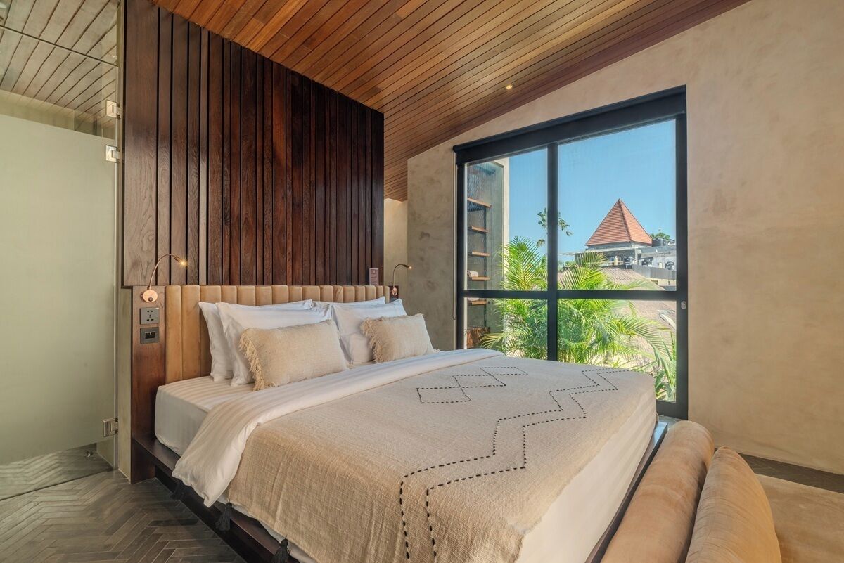 A modern bedroom with a wooden headboard, a large window with a view of palm trees and a building, and a bed with a geometric-patterned bedspread.