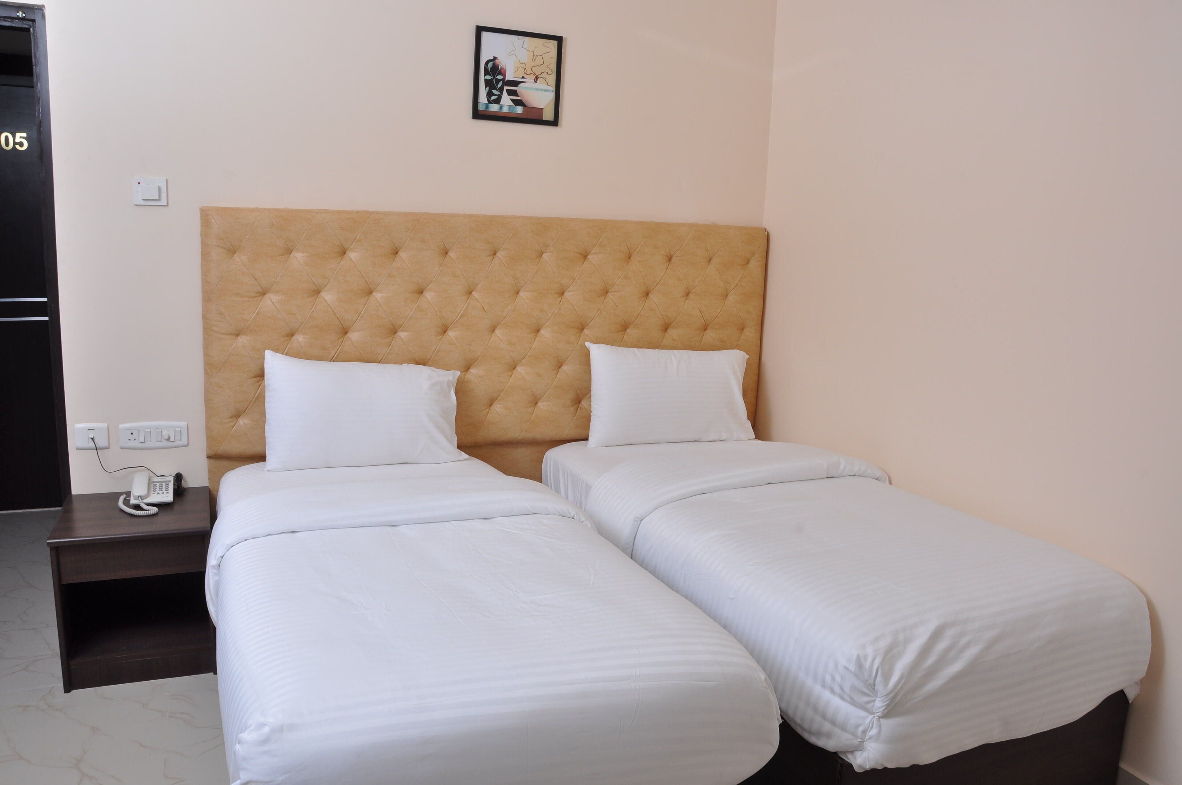 A hotel room with two single beds, a wooden headboard, a bedside table with a phone, and a wall-mounted light switch.