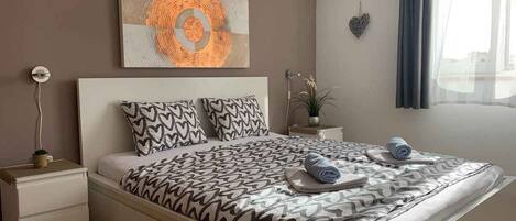 Un dormitorio moderno con cama, mesita de noche, un cuadro en la pared y una ventana con cortinas.