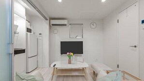 A compact room with a bed, a small table with a vase of flowers, a mounted TV, and a wall clock. - ANON Shibuya A103 (Tokyo)