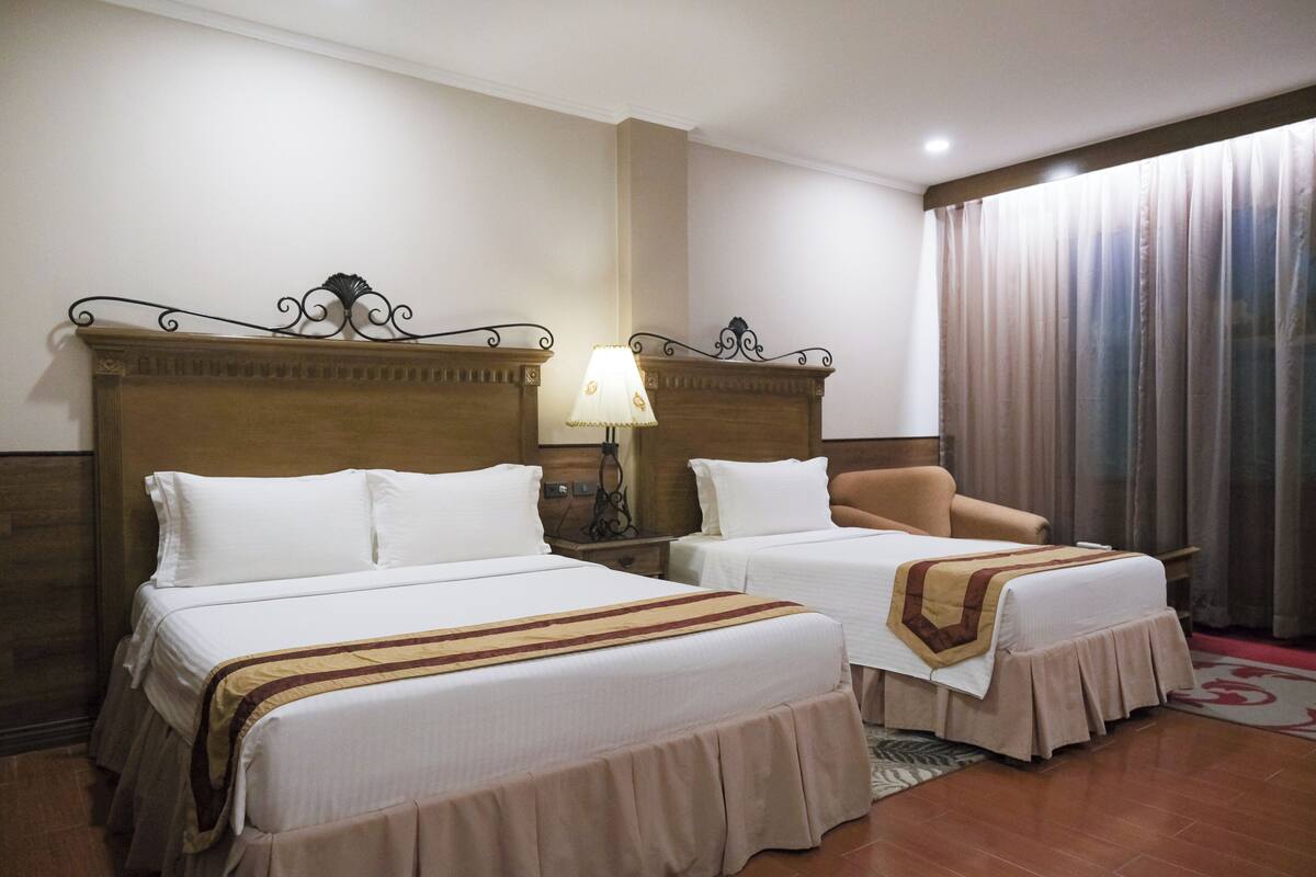 Una habitación de hotel con dos camas, una silla, una lámpara y un ventanal con cortinas.