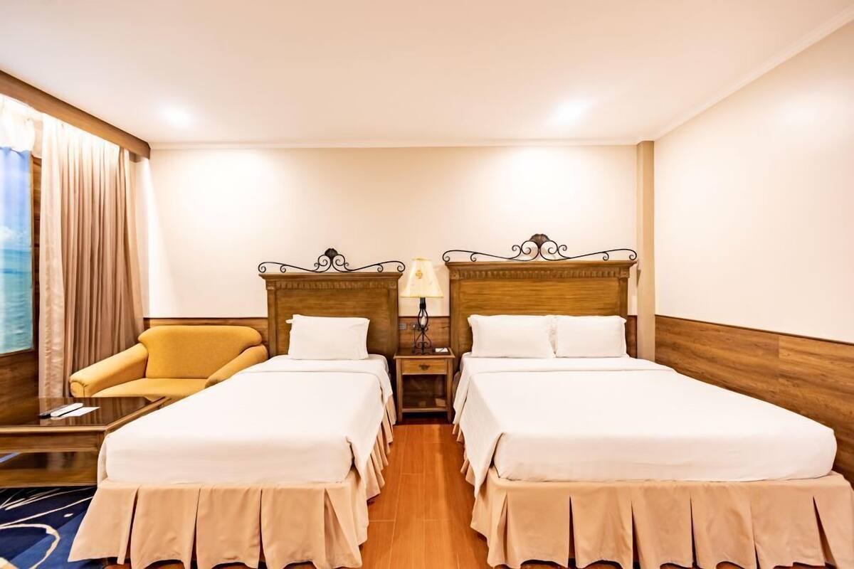 Habitación de hotel con dos camas, una silla, una lámpara y vista al mar.