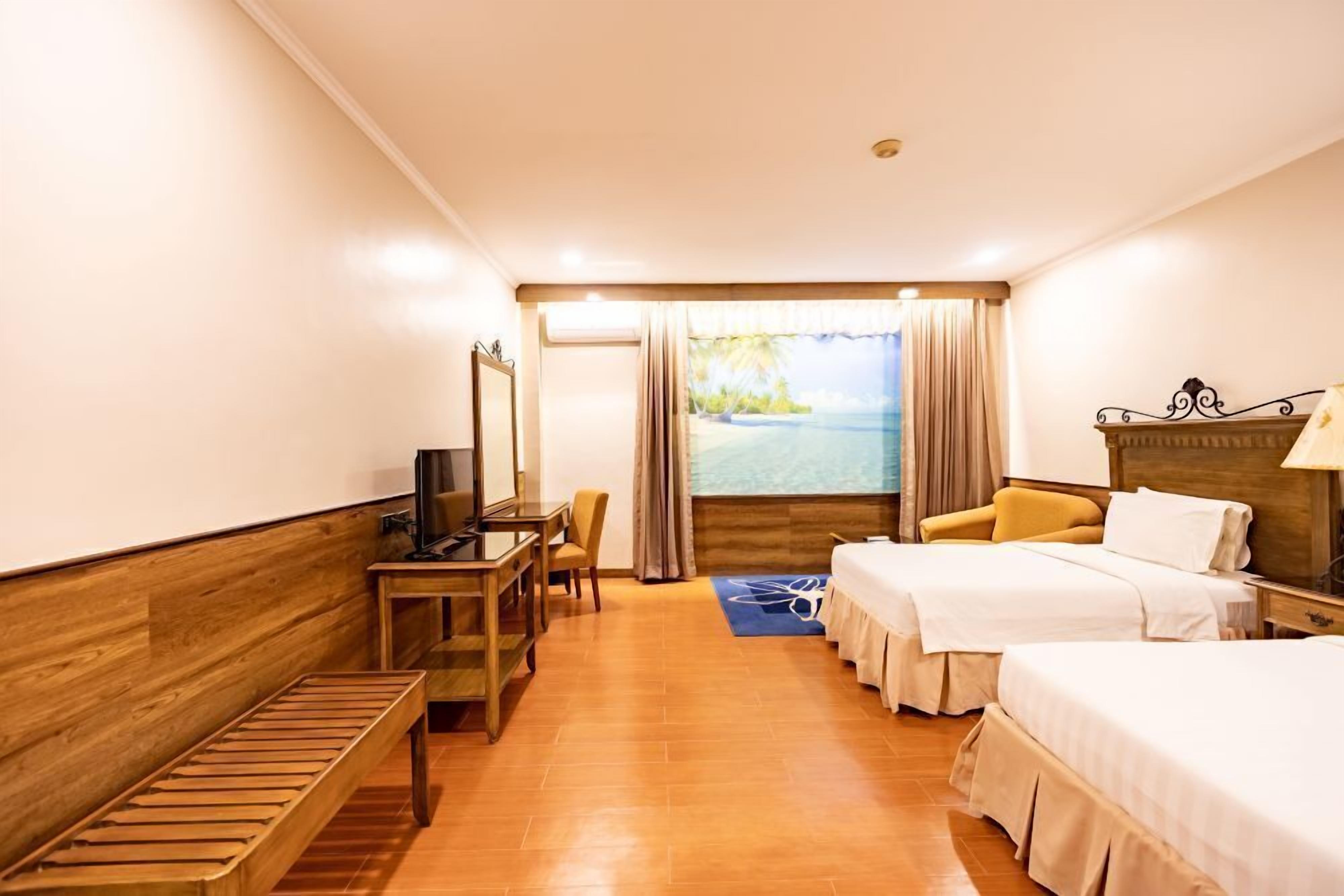 A hotel room with two beds, a desk, a chair, a TV, and a window with a view of a beach.