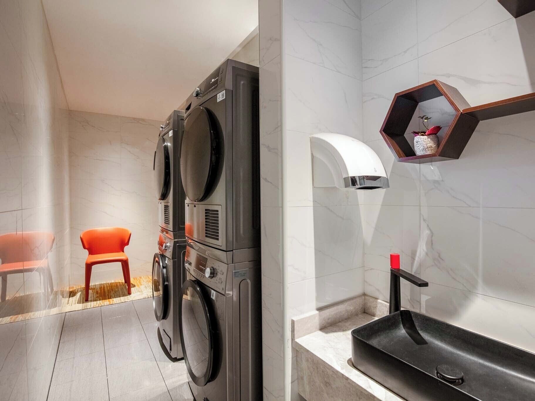 A modern laundry room with stacked washers and dryers, a small stool, and a sink with a wooden shelf above it.