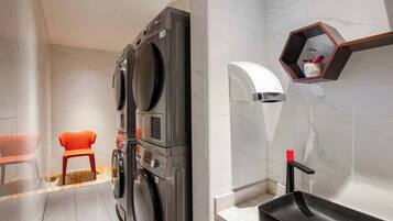 A modern laundry room with stacked washers and dryers, a small stool, and a sink with a wooden shelf above it.