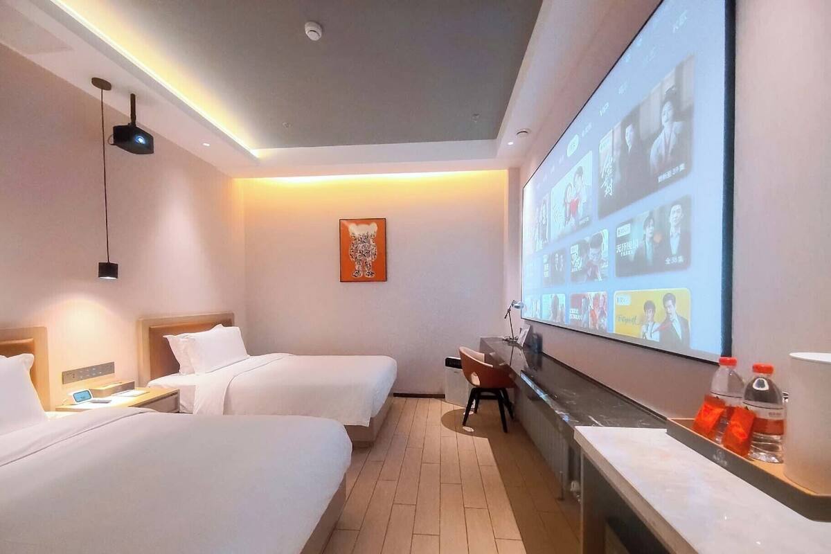 A hotel room with two beds, a desk, a large screen displaying a presentation, and a small table with a bottle and glasses.