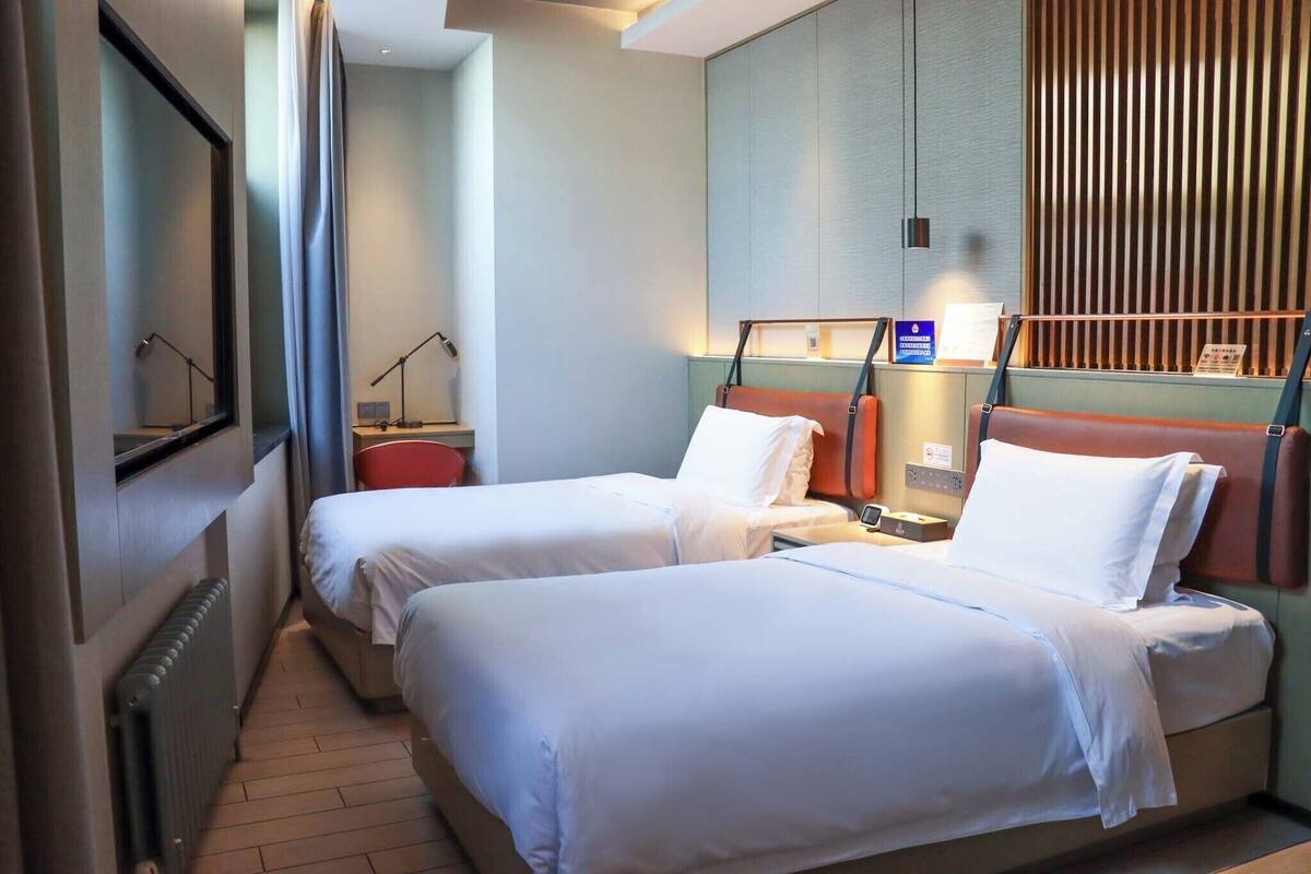A hotel room with two single beds, a nightstand, a lamp, a window with blinds, and a wall-mounted panel.