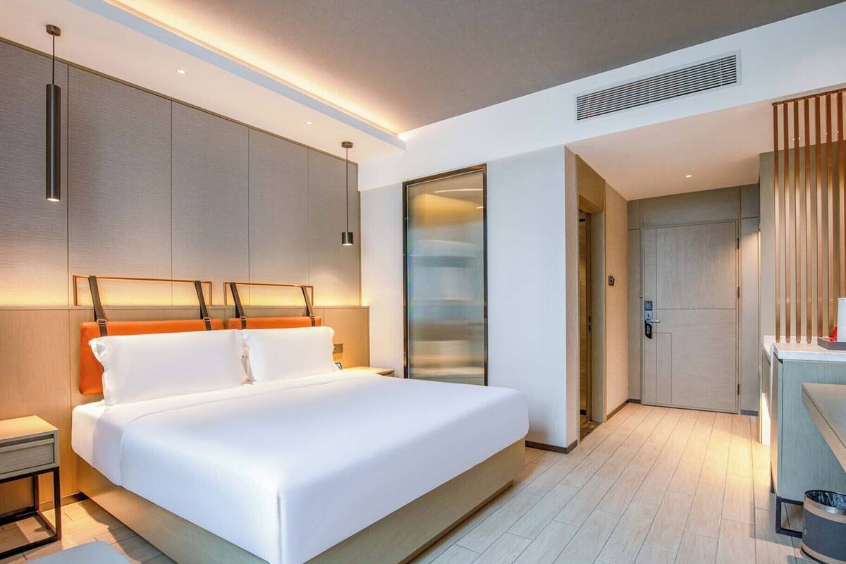A modern hotel room with a large bed, a bedside table, a nightstand, a built-in wardrobe, and a bathroom with a shower and toilet.