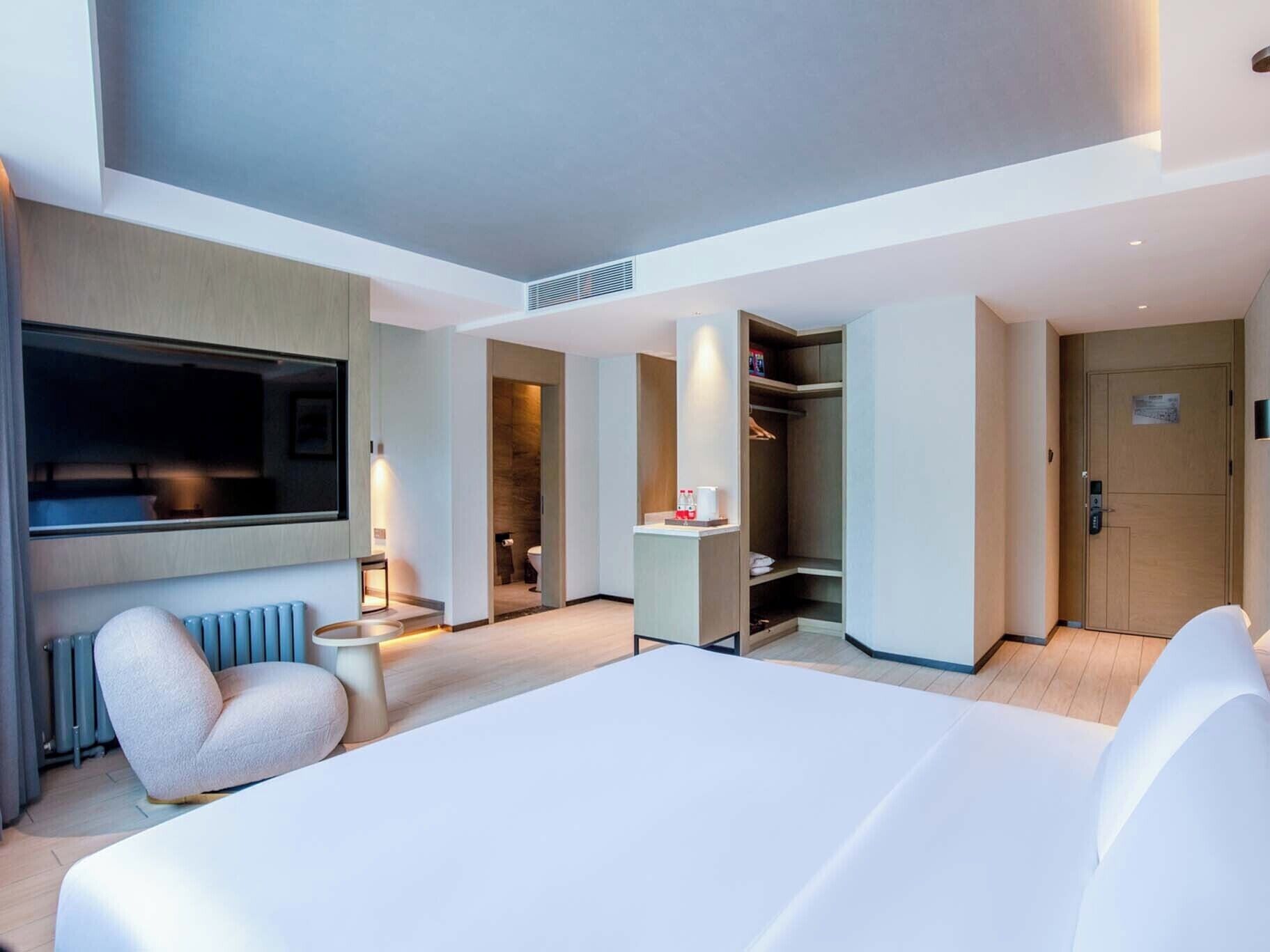 A modern hotel room with a large bed, a seating area with a chair, a small table, a TV mounted on the wall, and a walk-in closet.