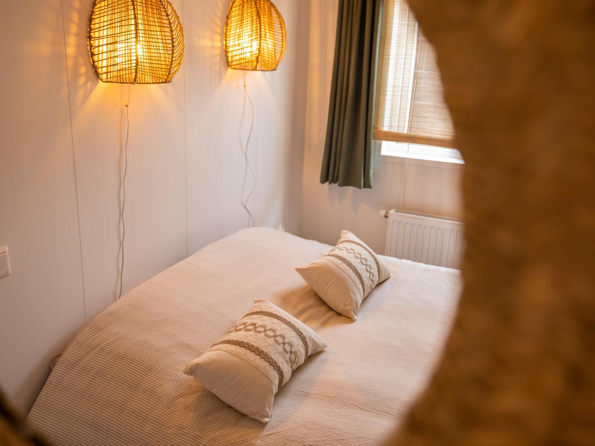 A bedroom with a bed, two wall-mounted lamps, a window with curtains, and a radiator.