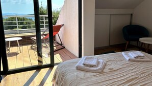 A bedroom with a large window, a bed with folded towels, a small table, and a chair.