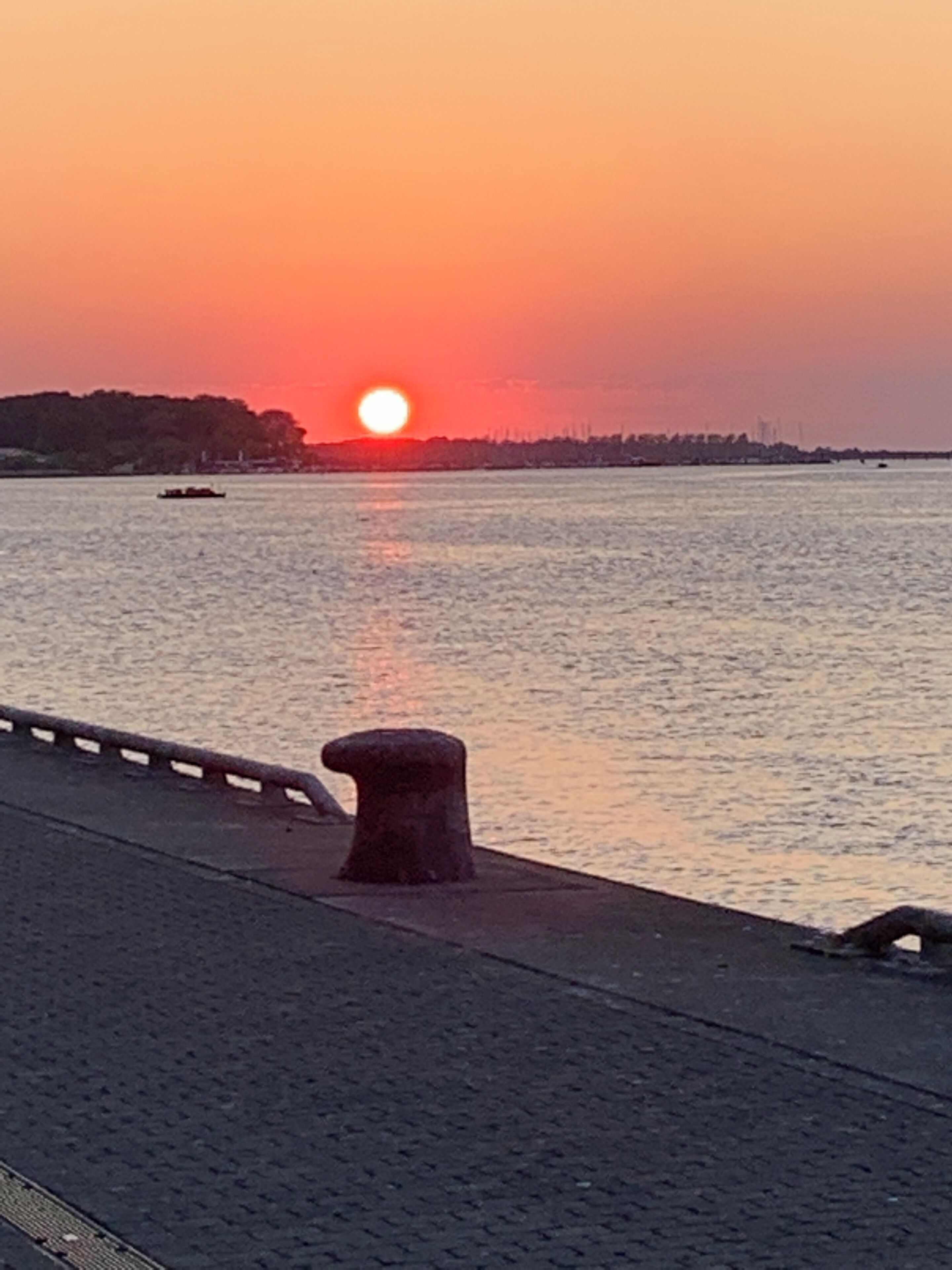 A sunset over a calm body of water with a distant shoreline and a bridge in the foreground.
