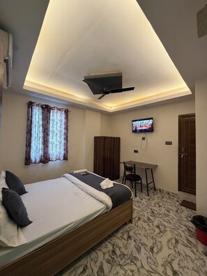 A bedroom with a bed, a desk, a TV, and a ceiling fan. - AV Grand Hotel (Kanpur)