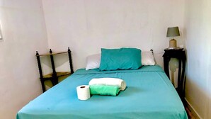 Una cama individual con una colcha color turquesa, una almohada verde y una almohada blanca. Hay una mesita de noche con una lámpara y una toalla enrollada sobre la cama.