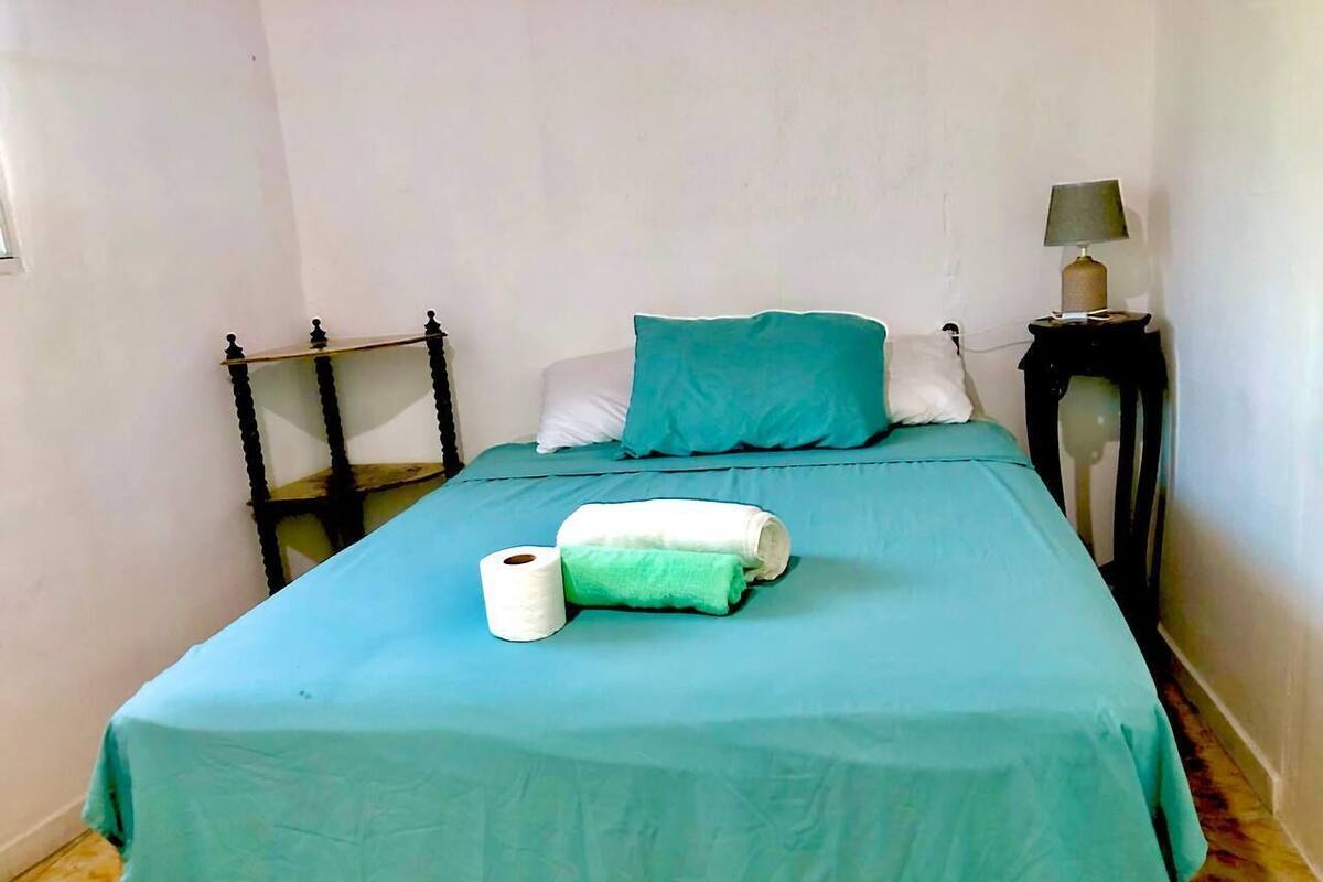 A single bed with a teal bedspread, a green pillow, and a white pillow. There is a small bedside table with a lamp and a rolled-up towel on the bed.