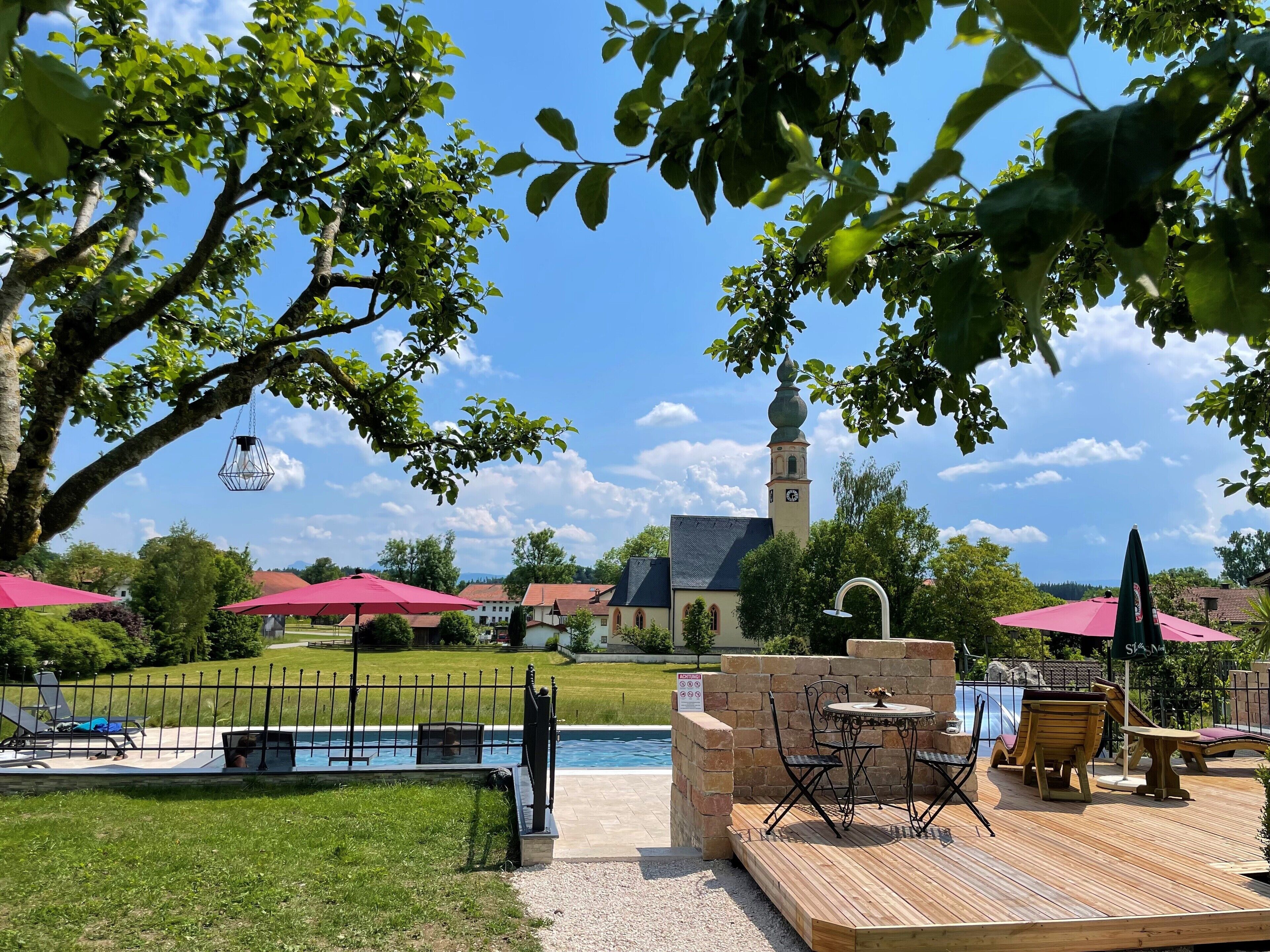 Un espace aménagé au bord de la piscine, avec une terrasse en bois, des sièges d’extérieur et une vue sur une église dotée d’un clocher.
