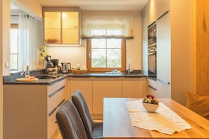 A compact kitchen with a dining area, featuring a coffee machine, a sink, and a window with a view of greenery. - Abendsonne Vacation Apartment, 60 m² (Palling)