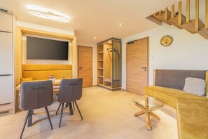 A modern living room with a sofa, a dining table with chairs, a TV mounted on the wall, and a wooden staircase. - Abendsonne Vacation Apartment, 60 m² (Palling)