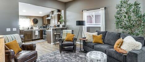 Een moderne woonkamer met een bank, fauteuil en poef, een keuken op de achtergrond, en een groot raam met gordijnen.