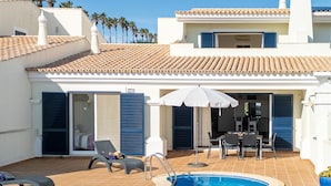 A swimming pool with lounge chairs and umbrellas, a dining area, and a building with a tiled roof and blue shutters.