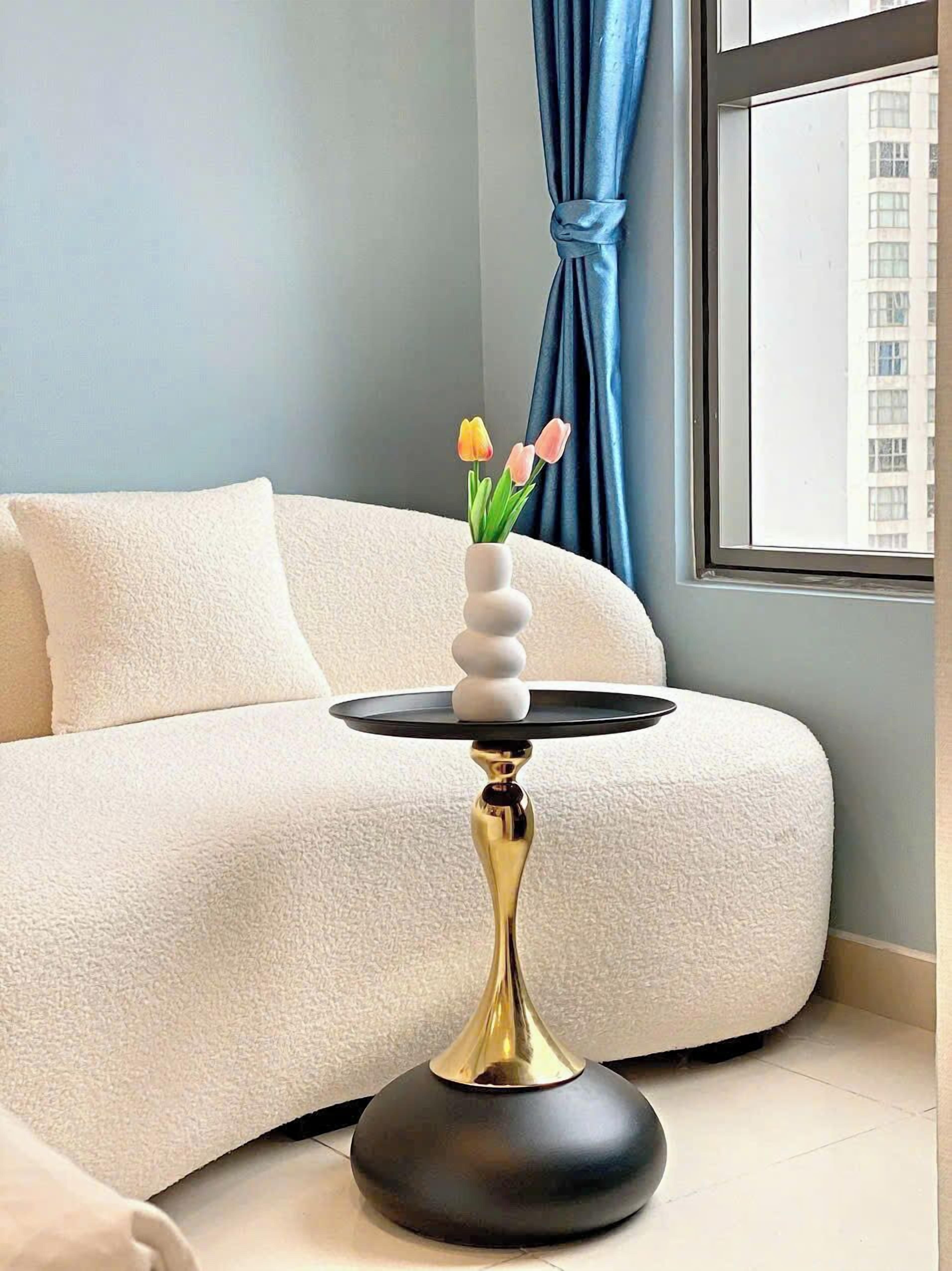 Un soggiorno moderno con un divano bianco, un tavolino nero e dorato con un vaso di tulipani, e vista su un edificio attraverso una finestra.