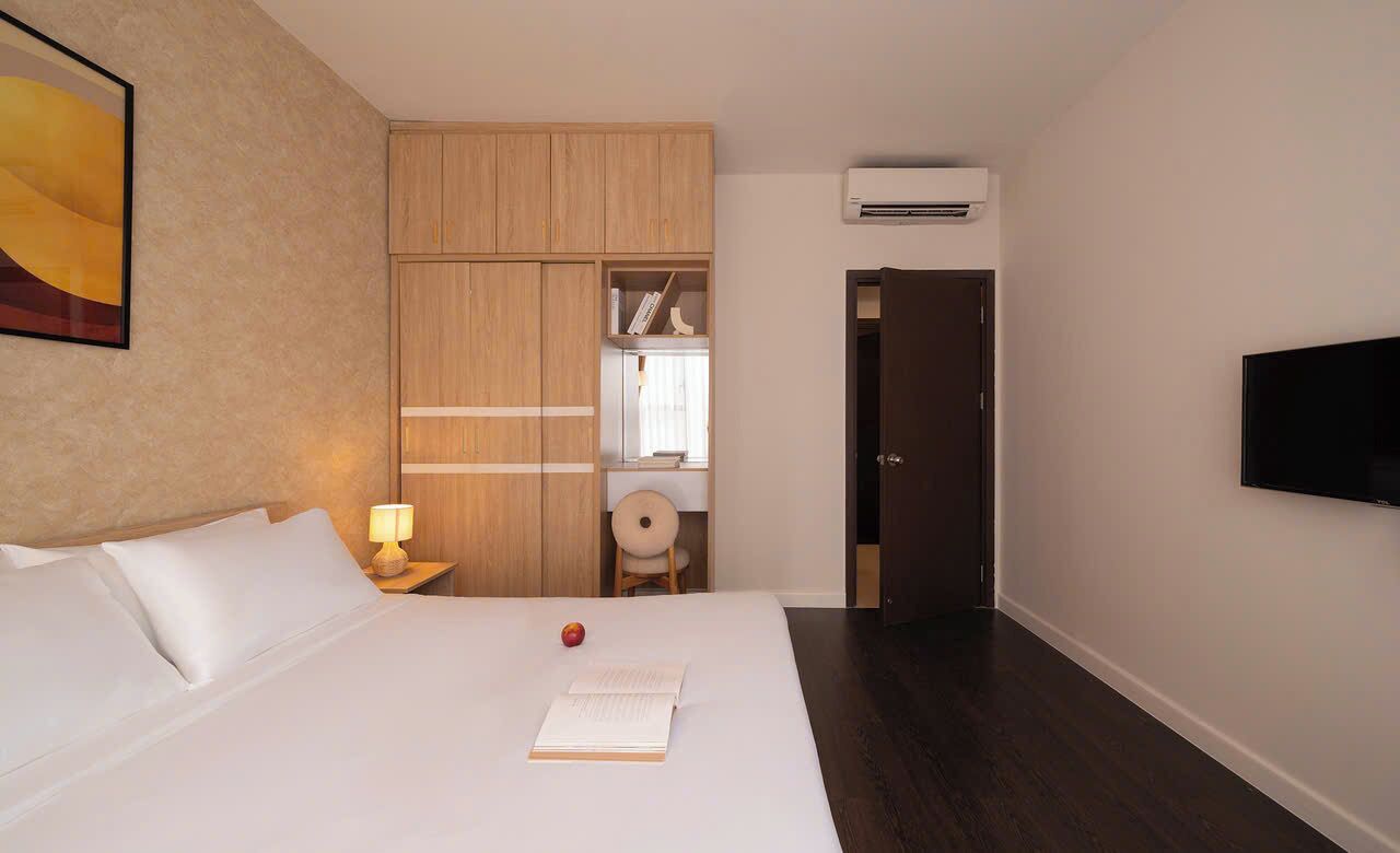 Ein Hotelzimmer mit einem Bett, einem Nachttisch, einer Lampe, einem Buch, Obst, einem Kleiderschrank, einem Fernseher und einer Klimaanlage.