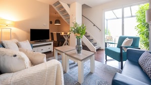 Una sala moderna con sofá, televisión, comedor, escalera y ventanas amplias.