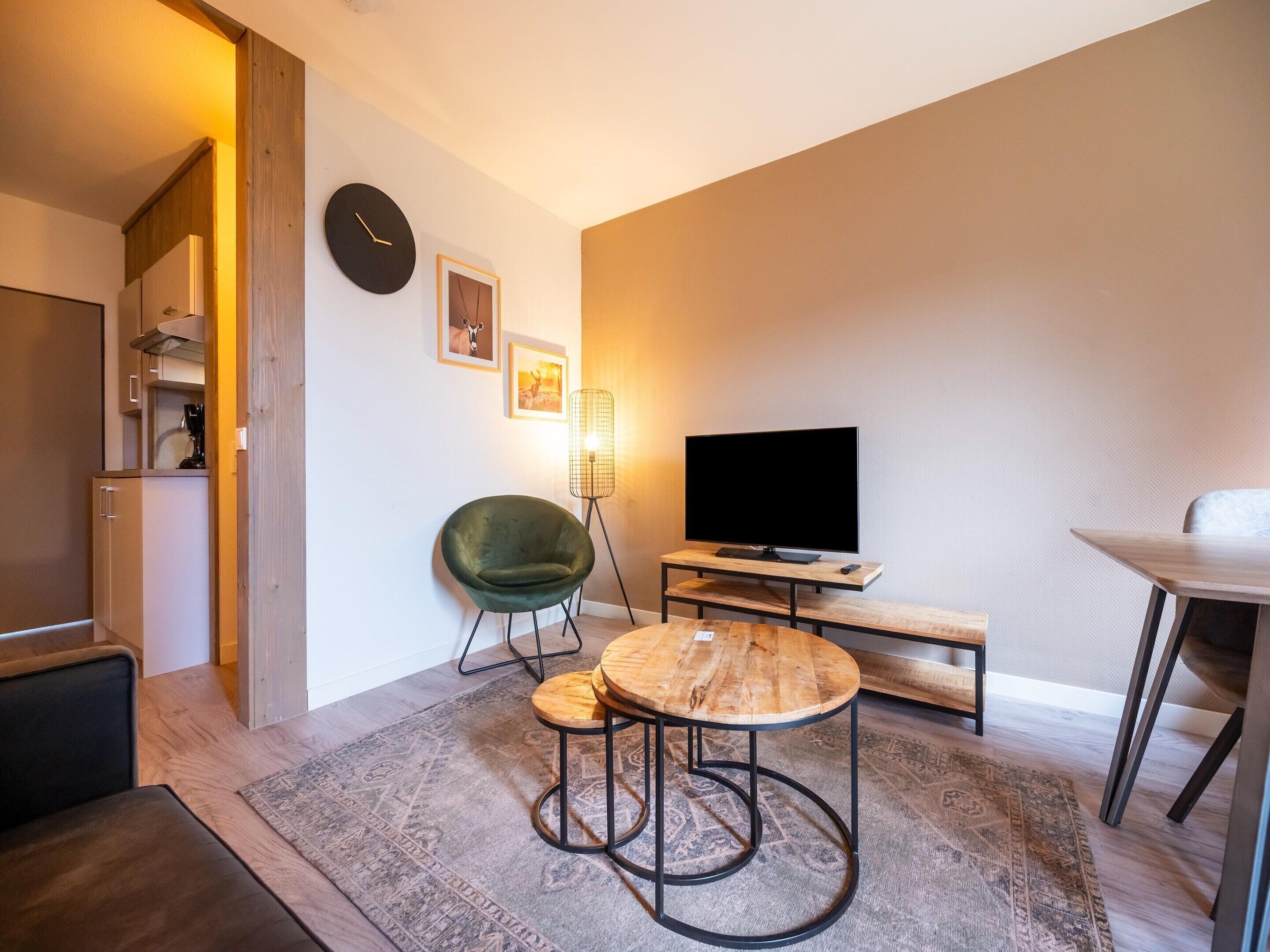 A modern living room with a flat-screen TV, a green chair, a wooden coffee table, and a dining area with a table and chairs.