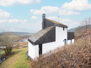 A small white building with a dark roof, surrounded by vegetation and a hilly landscape.