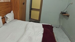 Un dormitorio con cabecera de madera, cama con ropa blanca, manta roja y una pequeña estantería con lámpara e interruptor.