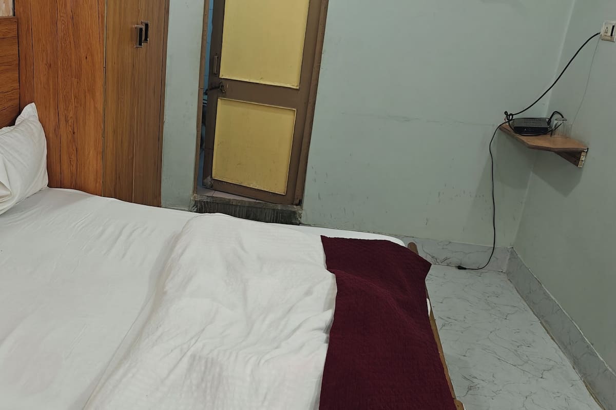 A bedroom with a wooden headboard, a bed with white bedding, a red throw blanket, and a small shelf with a lamp and power outlet.
