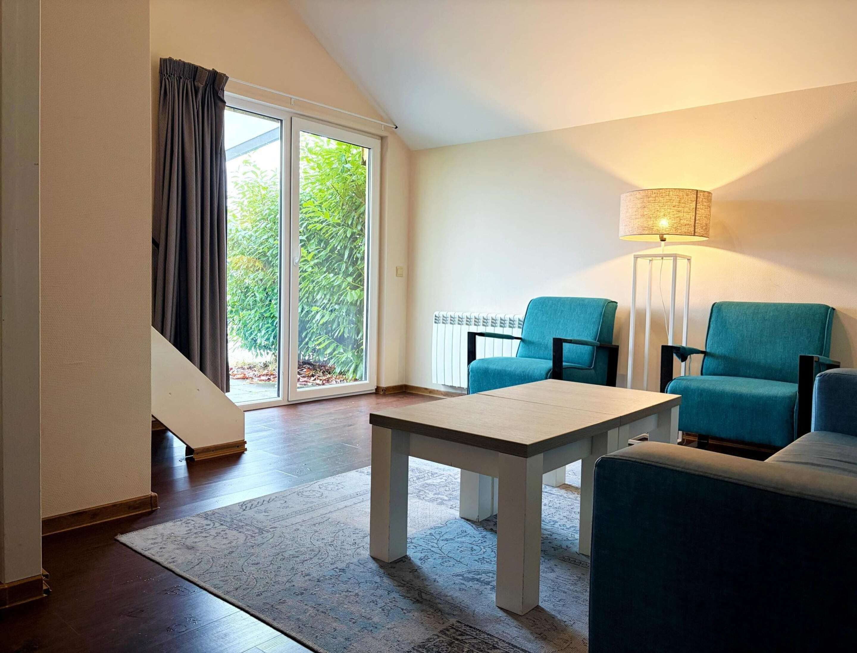 Un salon moderne avec un canapé, une table et une lampe. Une porte-fenêtre coulissante donne sur un balcon agrémenté de plantes.