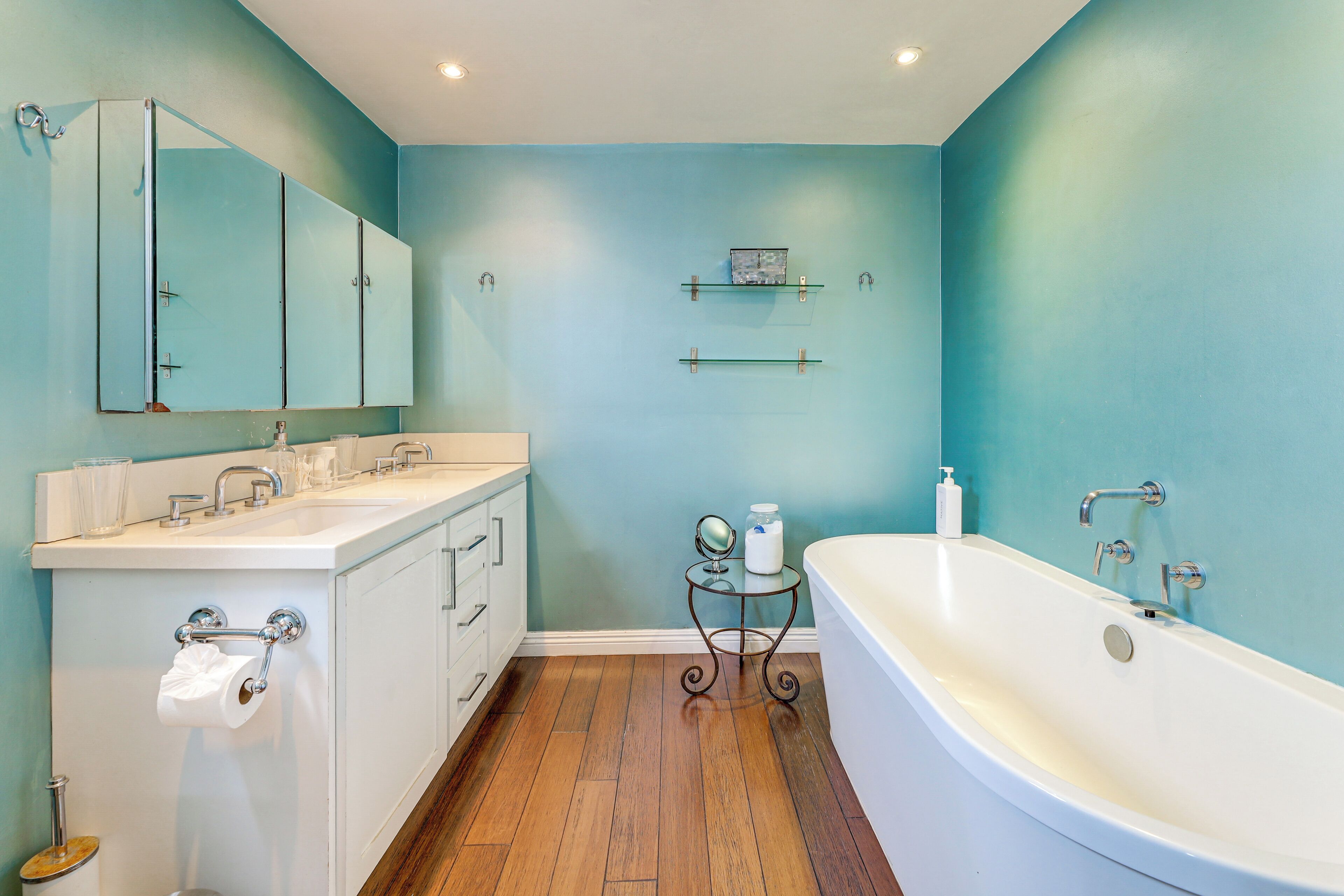 A bathroom with a white bathtub, a sink with a mirror above it, and a toilet. There are shelves on the wall and a small table with a mirror and a bottle.